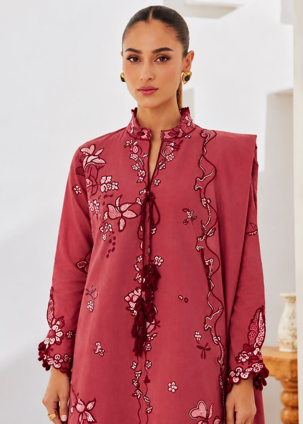 A woman wearing a dusty rose pink embroidered tunic with floral and vine patterns, featuring a high collar, tassel tie, and sleeve cuffs, set against a bright indoor background.