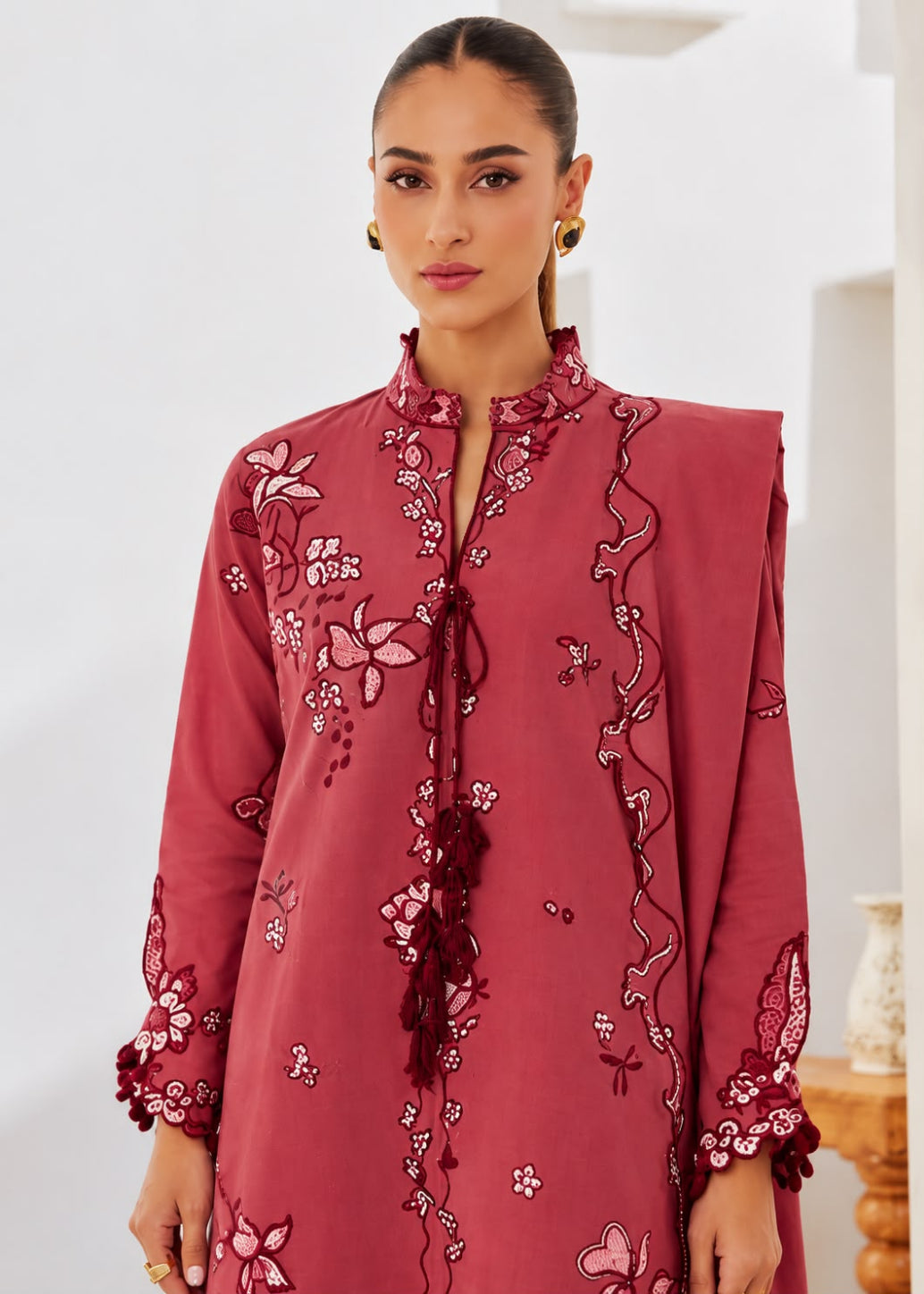 A woman wearing a dusty rose pink embroidered tunic with floral and vine patterns, featuring a high collar, tassel tie, and sleeve cuffs, set against a bright indoor background.