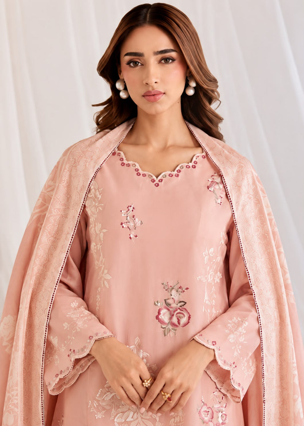 A woman modeling a light pink traditional outfit with floral embroidery and a matching draped scarf, photographed against a soft white curtain backdrop.