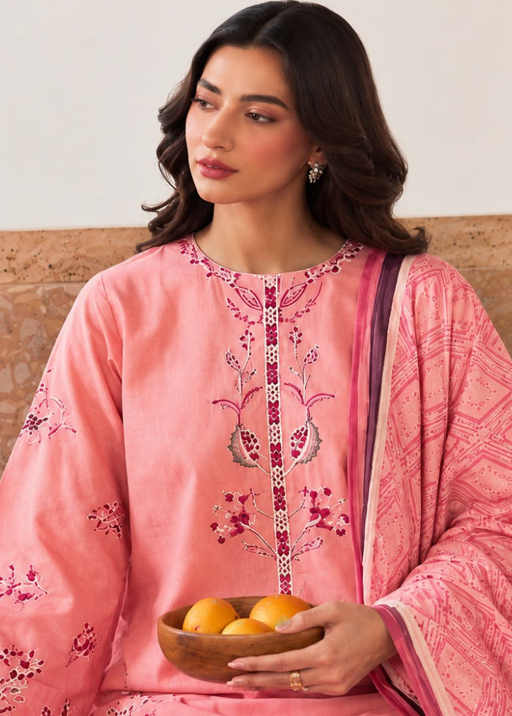 A woman wearing a pink embroidered traditional outfit with matching dupatta, seated indoors and holding a small bowl of oranges.