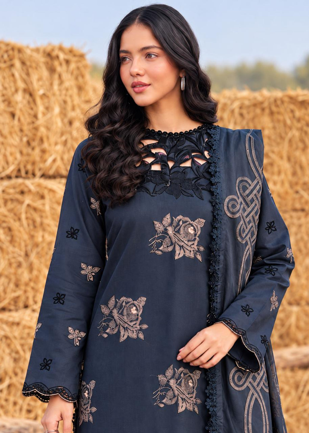 A woman wearing a navy blue embroidered kurta set with a matching dupatta, standing outdoors in front of a field of golden hay bales. The kurta features intricate floral embroidery and lace detailing.