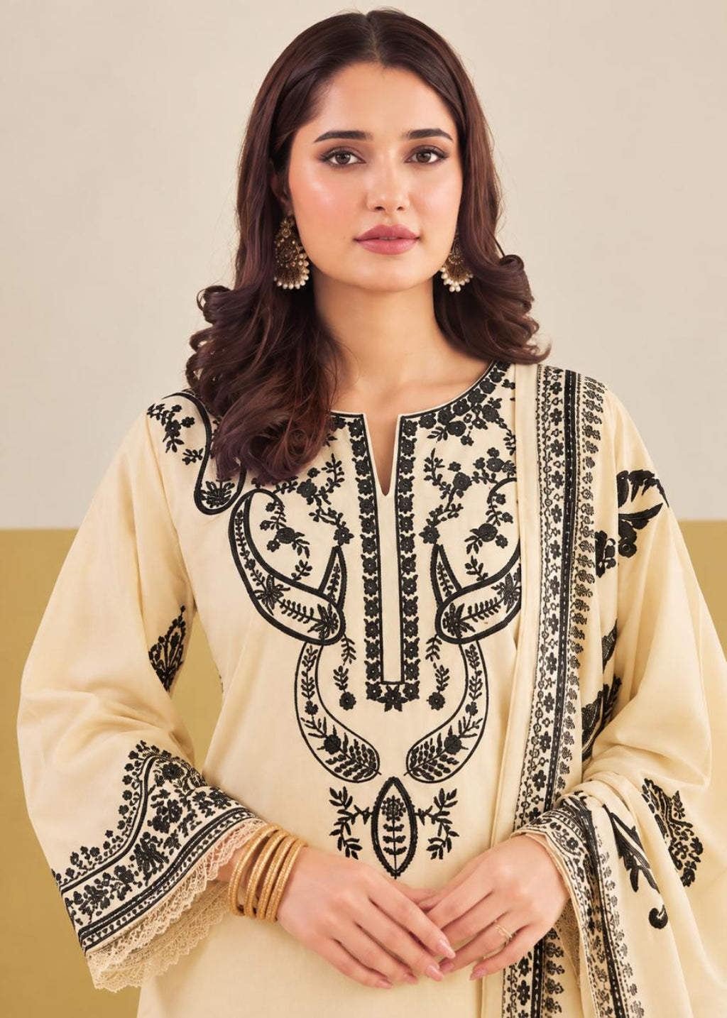 A woman wearing a cream-colored traditional embroidered kurta with black floral embroidery and a matching embroidered dupatta, styled with gold bangles and earrings.
