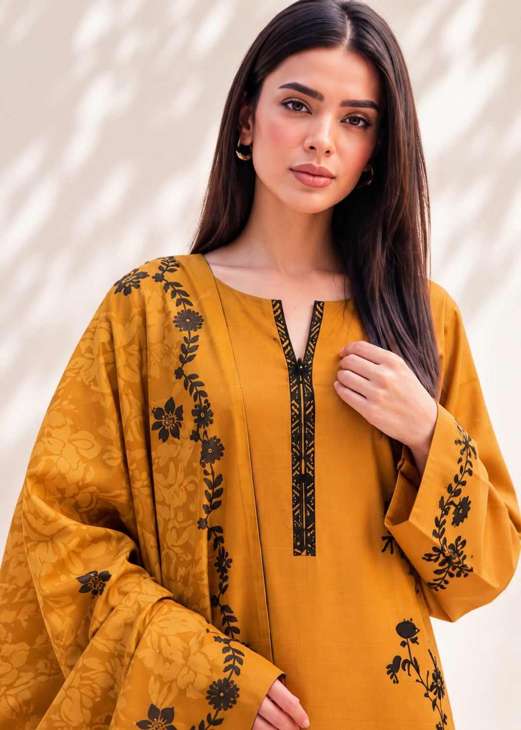 A woman modeling a mustard yellow embroidered kurta with black floral and geometric embroidery details, set against a light beige background with soft leaf shadows.