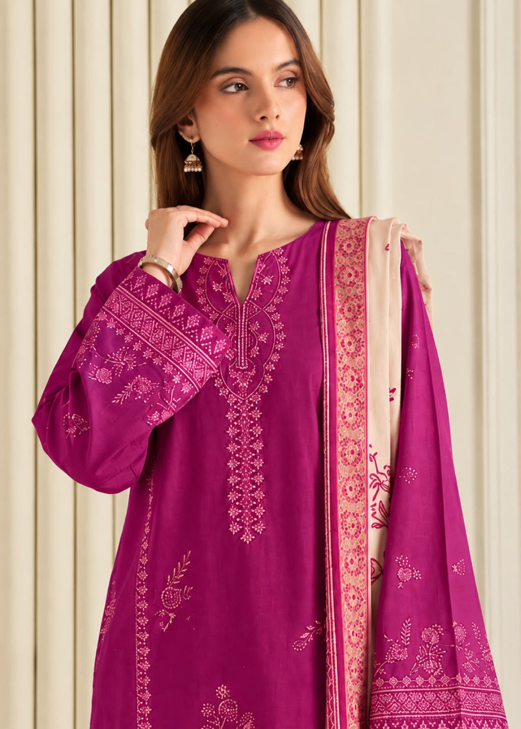 A woman wearing a magenta pink embroidered traditional kurta with a matching dupatta, set against a neutral cream-colored paneled background.
