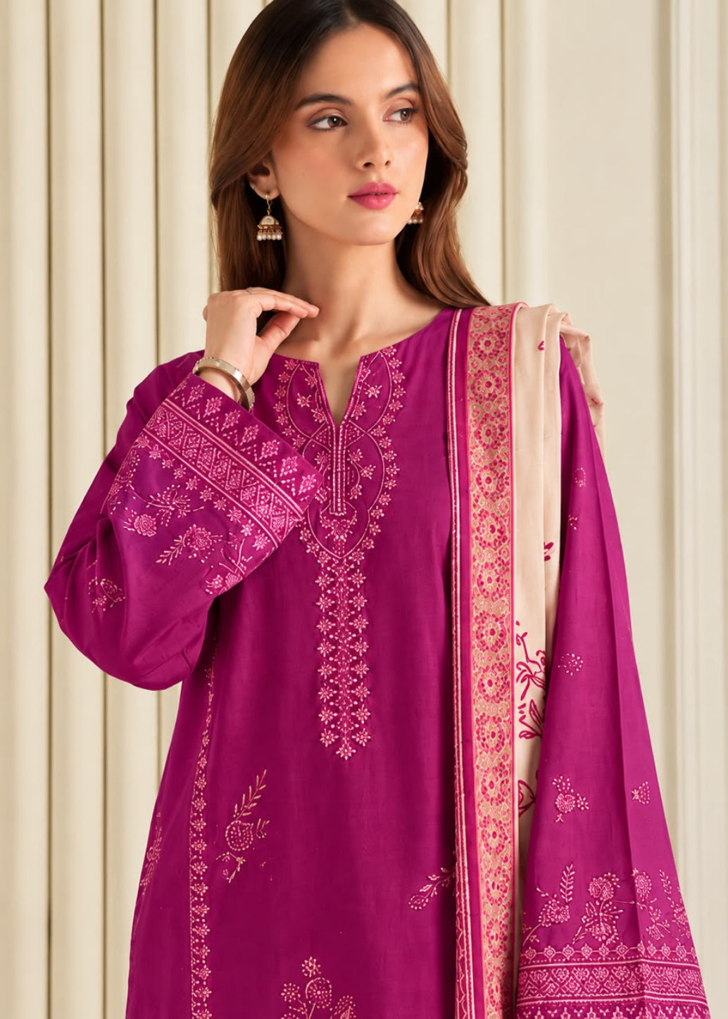 A woman wearing a magenta pink embroidered traditional kurta with a matching dupatta, set against a neutral cream-colored paneled background.