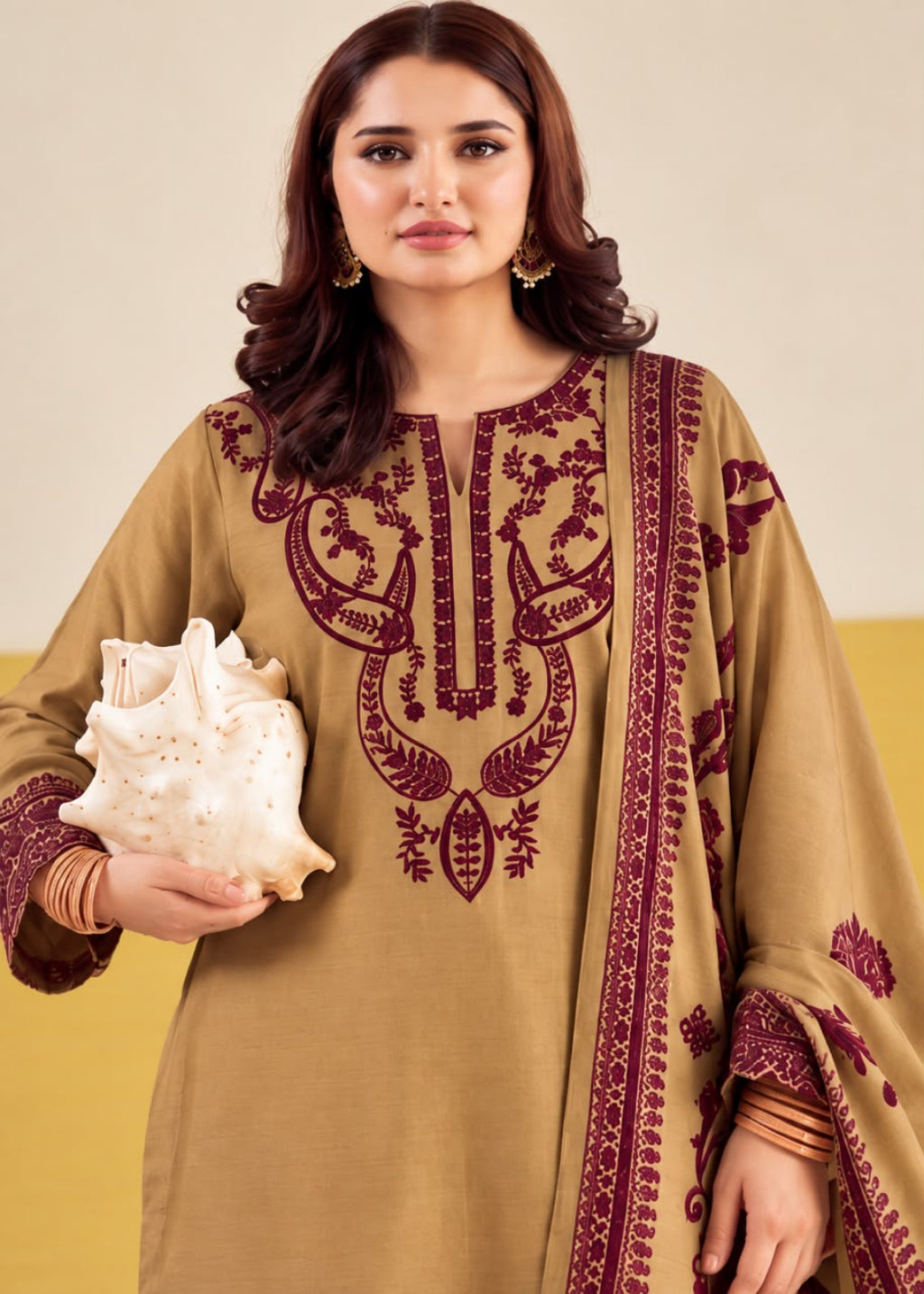 A woman modeling a beige kurta with deep maroon embroidery and a matching embroidered dupatta, holding a white seashell against a neutral studio background.