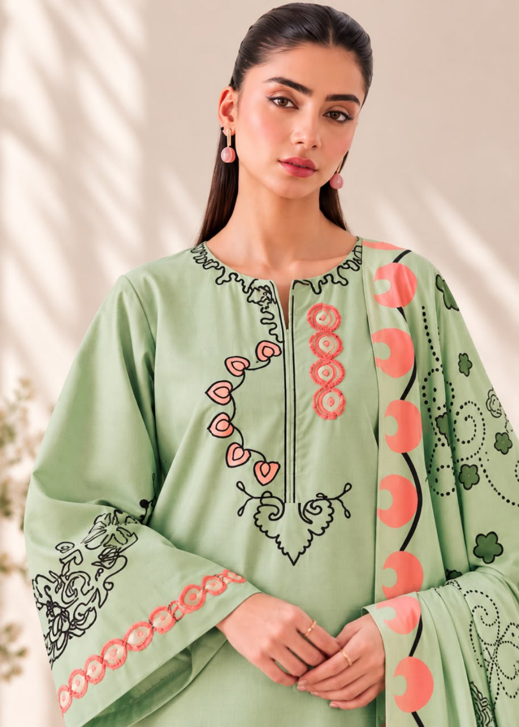 A woman wearing a light sage green embroidered tunic with pink and black details, set against a softly lit beige background with leaf shadows.