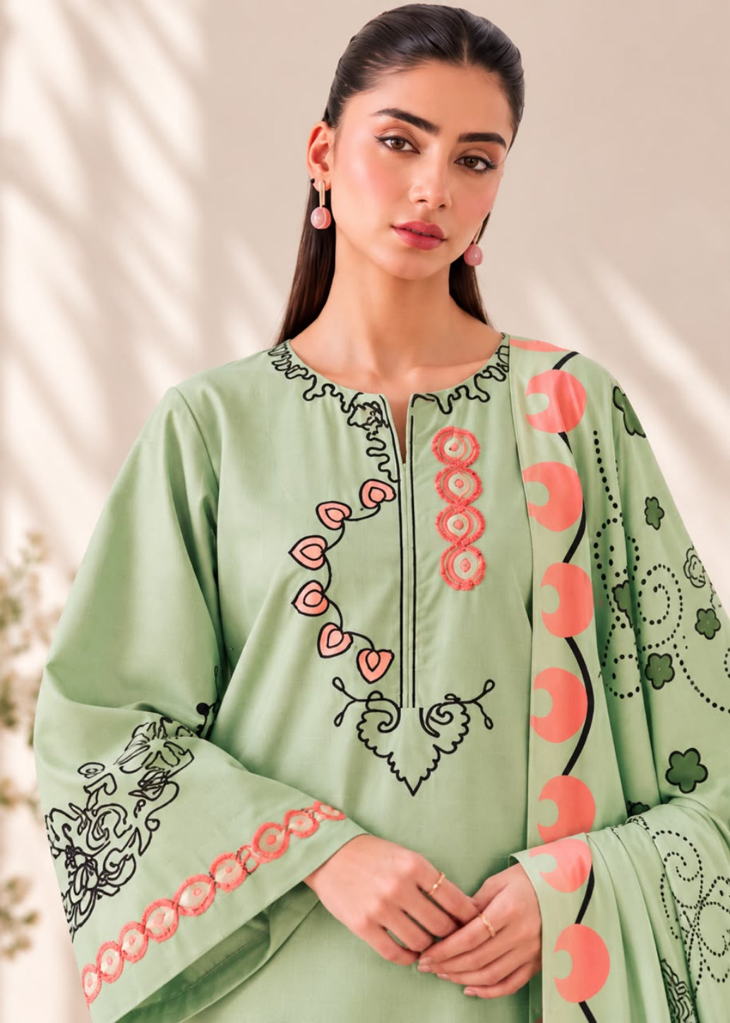 A woman wearing a light sage green embroidered tunic with pink and black details, set against a softly lit beige background with leaf shadows.