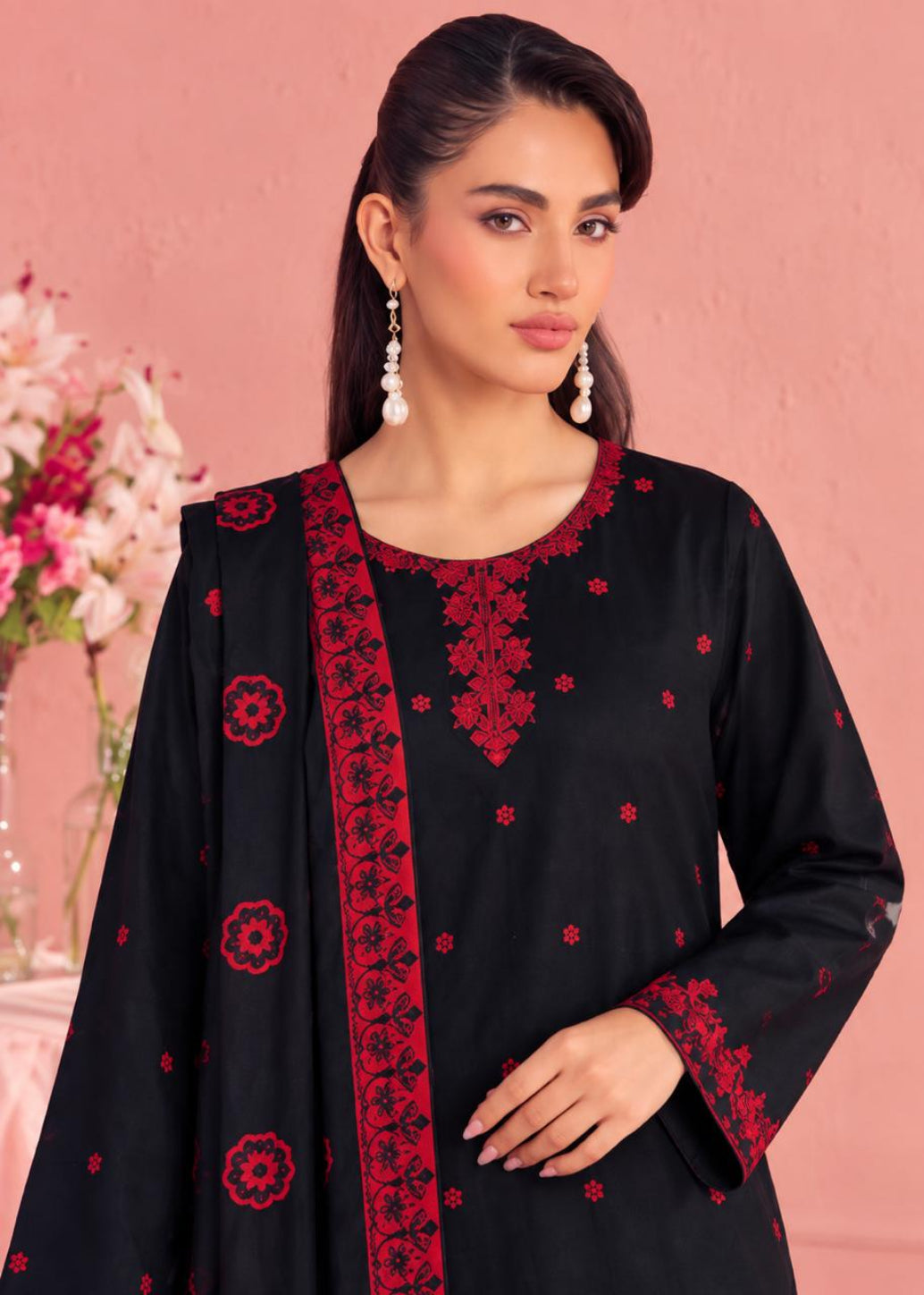 A woman modeling a black traditional outfit with red floral embroidery and a red border, posed against a pink wall with flowers in the background.