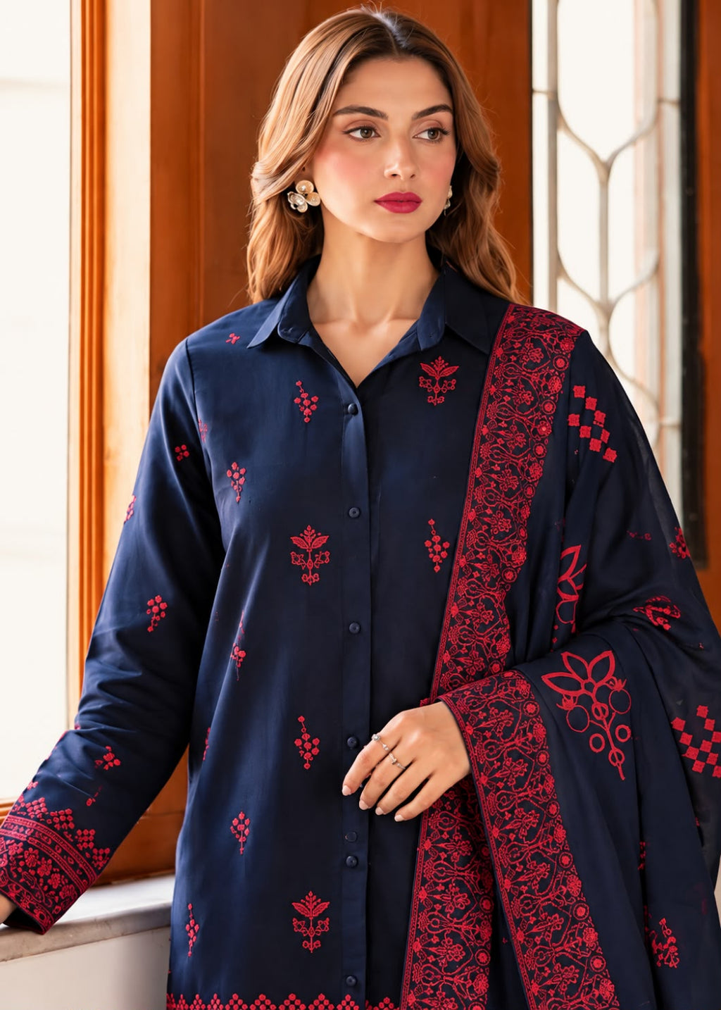 A woman wearing a navy blue kurta with red floral embroidery and a matching red border dupatta, posed in front of a warm-toned window.