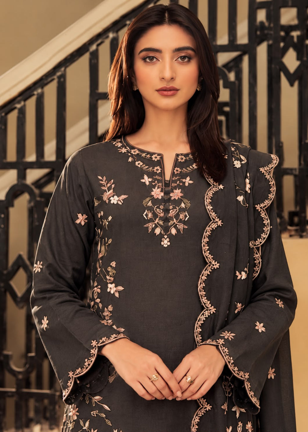 A woman wearing a dark charcoal gray embroidered kurta with floral patterns and scalloped borders, paired with a matching dupatta, posed in front of an indoor staircase.