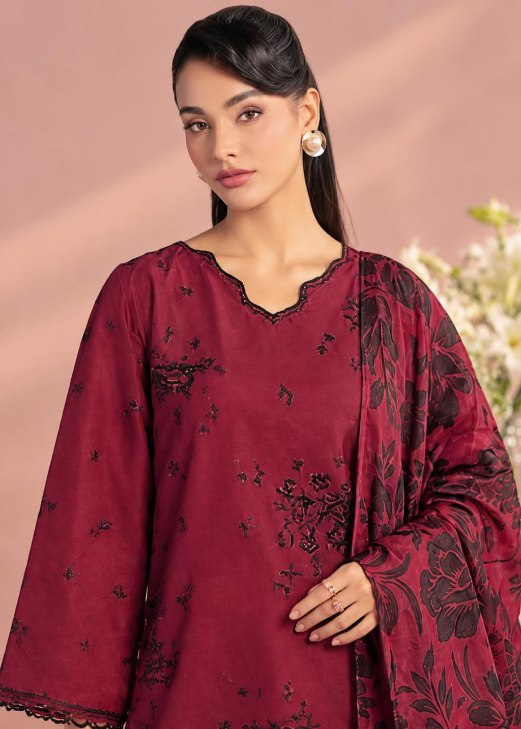 A woman modeling a deep maroon embroidered kurta with wide sleeves and a matching dupatta, featuring black floral embroidery and a scalloped neckline, set against a soft pink background.