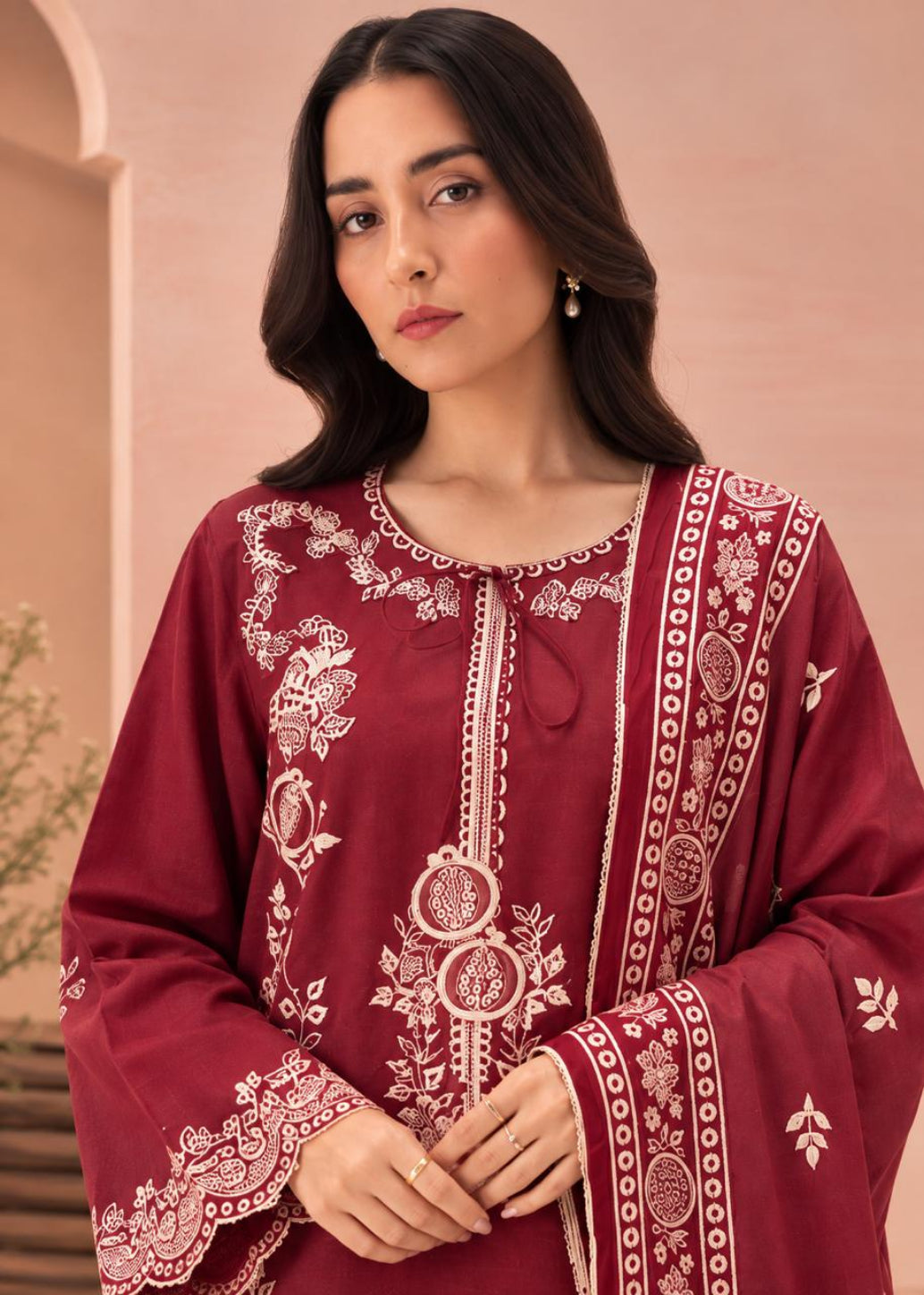 A woman wearing a deep maroon long-sleeve kurta with intricate white embroidery, paired with a matching embroidered dupatta, set against a soft peach-colored architectural background.