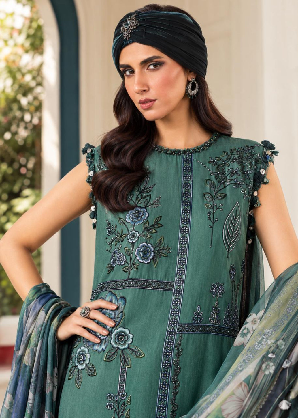 A woman models a green embroidered traditional outfit with floral detailing, a matching dupatta, and a black headwrap in an outdoor architectural setting.