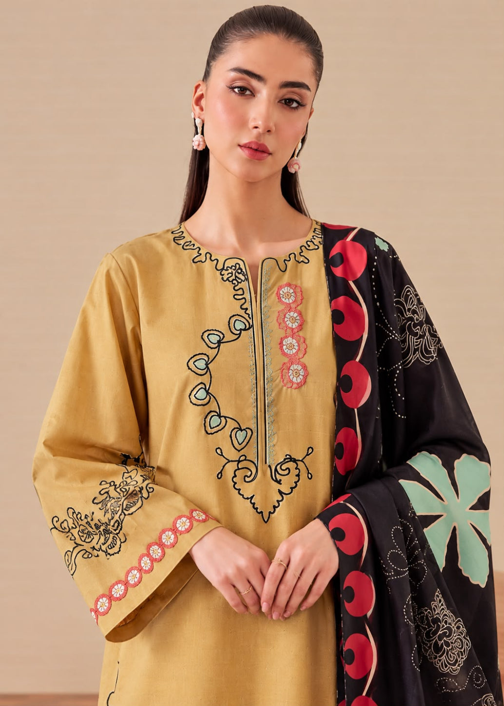 A woman modeling a mustard yellow embroidered kurta with a black dupatta featuring red and mint green floral patterns, set against a plain beige background.