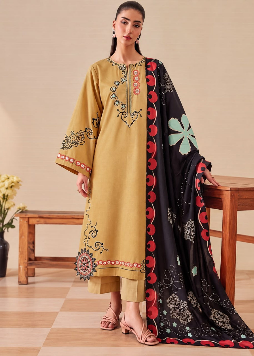 A woman models a traditional Indian salwar kameez suit set. The kurta is mustard yellow with colorful embroidery on the chest and sleeves, paired with matching trousers and a black dupatta featuring red scalloped borders and green and blue floral motifs.