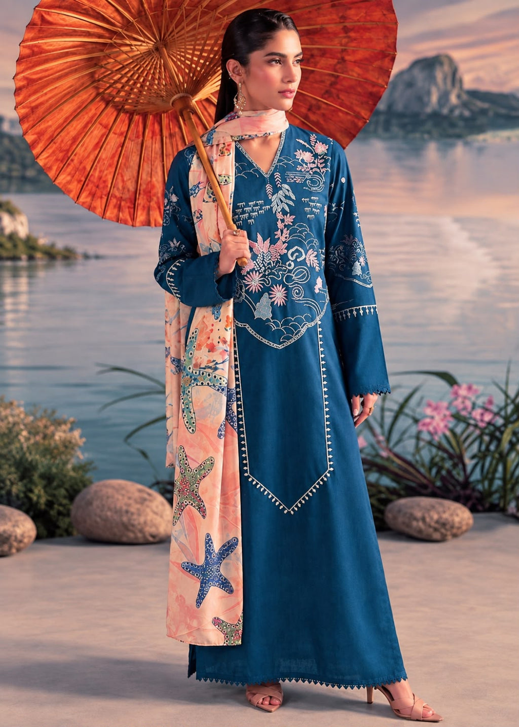 A woman stands outdoors by a waterfront holding a large orange parasol, wearing a long teal-blue embroidered kurta with a matching scarf featuring starfish and floral prints.