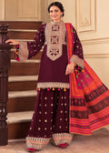 A woman models a traditional Indian salwar kameez suit in deep maroon with intricate gold and silver embroidery, paired with a bright pink and orange patterned dupatta, standing in front of a beige wall and wooden staircase.