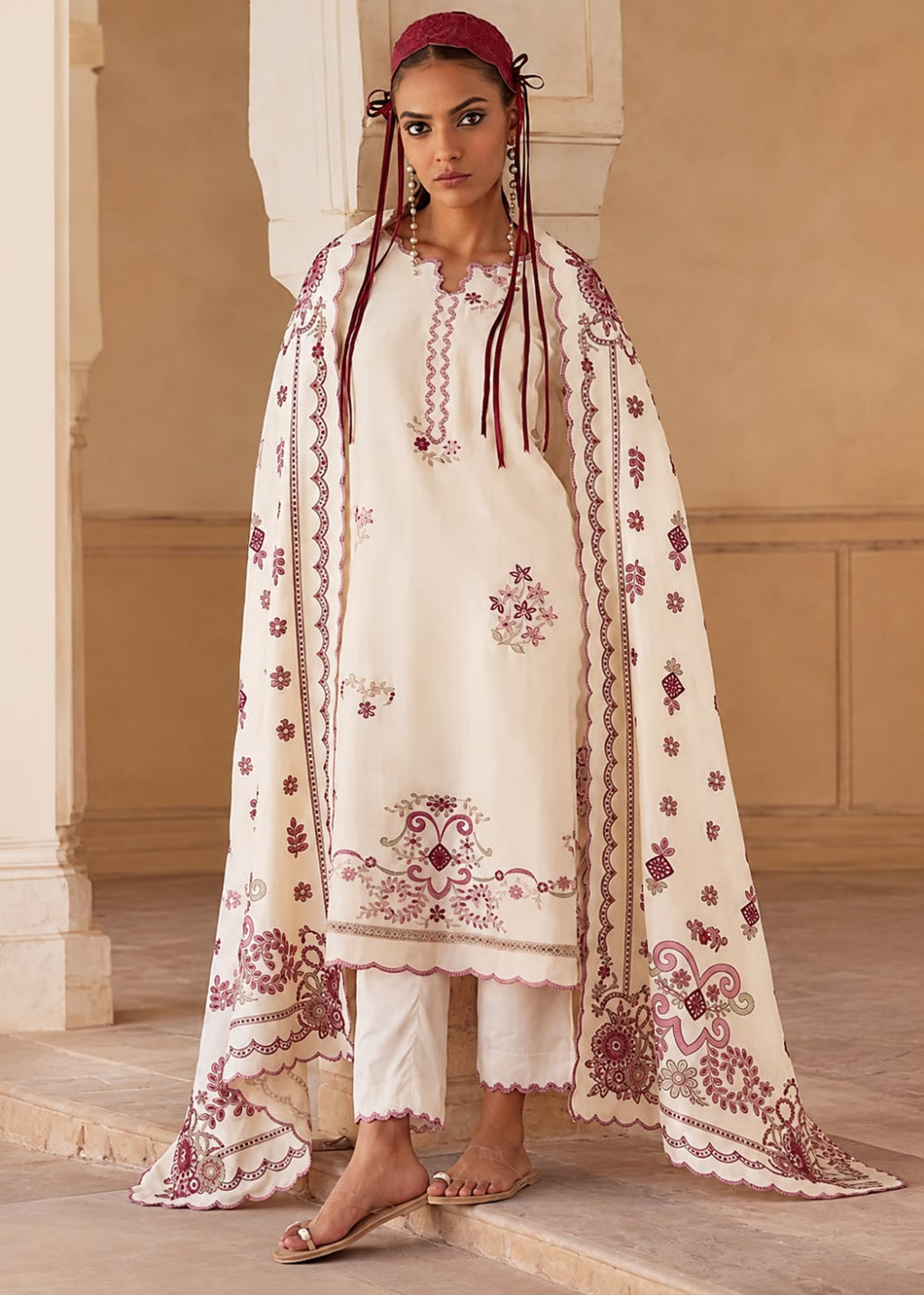 A woman models a traditional South Asian outfit consisting of an off-white embroidered kurta, matching white pants, and a large off-white dupatta with burgundy floral embroidery, set against a warm beige architectural background.