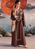 A woman stands outdoors by a water feature holding a large orange parasol, wearing a long brown embroidered dress with a floral scarf.