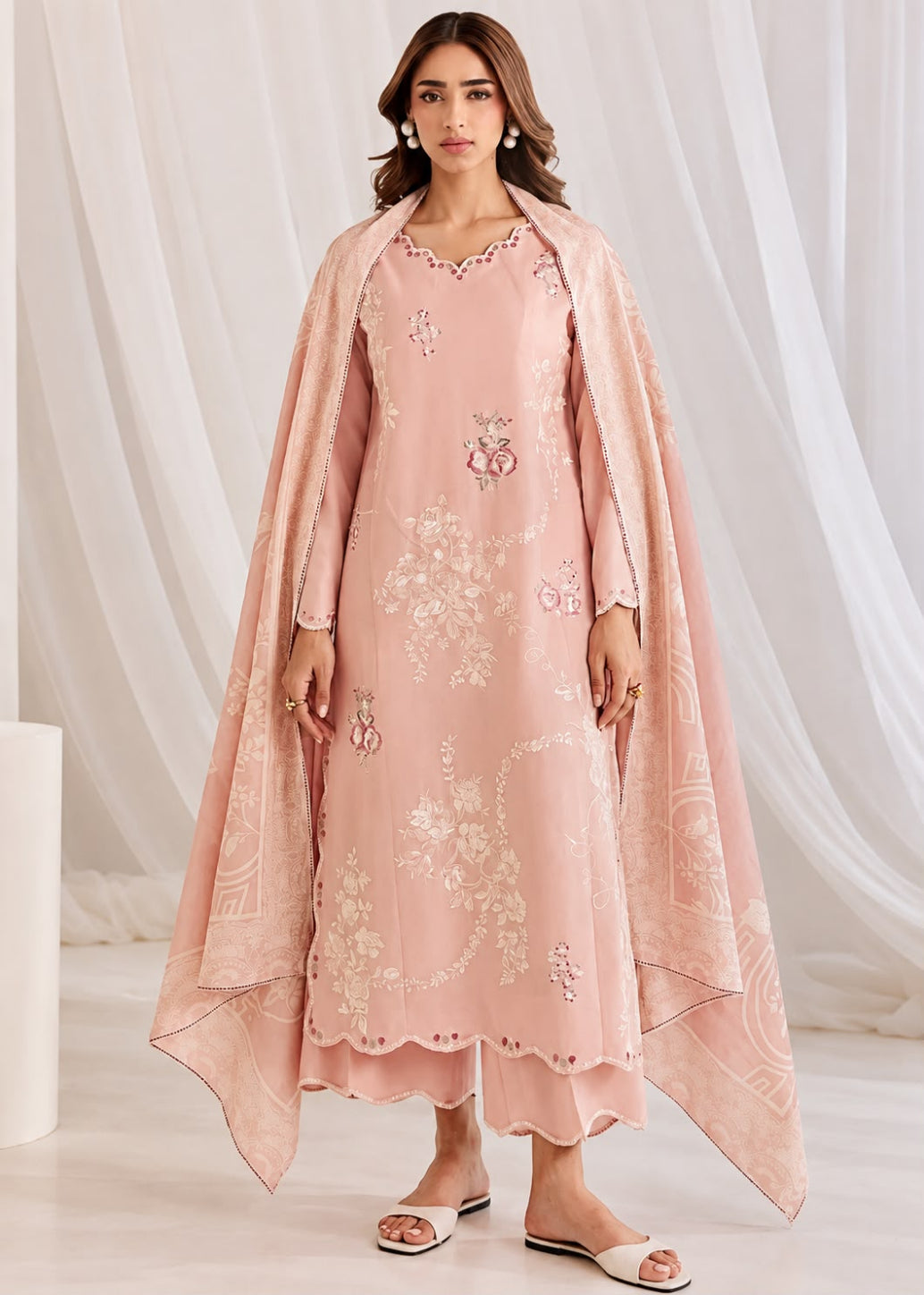A woman models a light pink traditional outfit consisting of a long kurta with floral embroidery, matching pants, and a sheer draped shawl, set against a soft white draped fabric background.