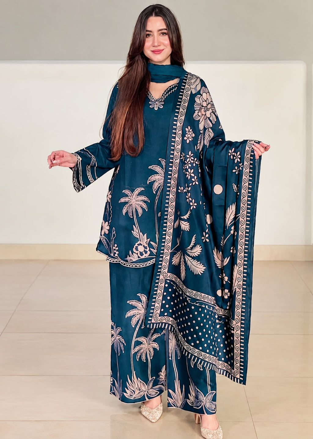 A woman modeling a dark teal blue three-piece traditional outfit with beige palm tree and floral prints, featuring a kurta, pants, and dupatta, set against a neutral indoor background.