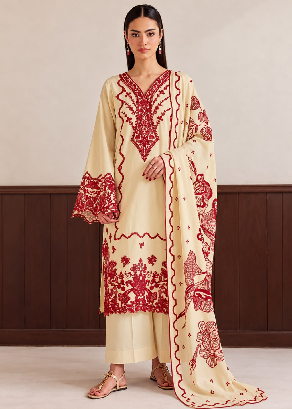A woman models a cream-colored three-piece ethnic suit with vibrant red floral embroidery. The set includes a long-sleeved kurta with a V-neck, matching trousers, and a dupatta featuring detailed red floral and paisley patterns.