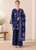 A woman models a navy blue three-piece traditional outfit with pink and white floral embroidery, consisting of a long-sleeve kurta, matching wide-leg trousers, and a dupatta, set against a warm beige background with a wooden bench and decorative vase.