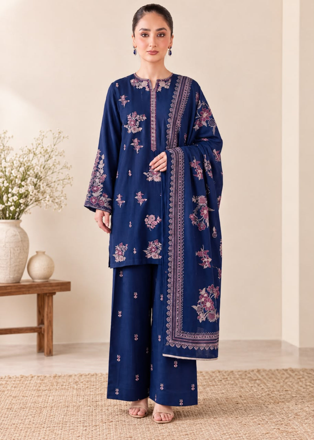 A woman models a navy blue three-piece traditional outfit with pink and white floral embroidery, consisting of a long-sleeve kurta, matching wide-leg trousers, and a dupatta, set against a warm beige background with a wooden bench and decorative vase.