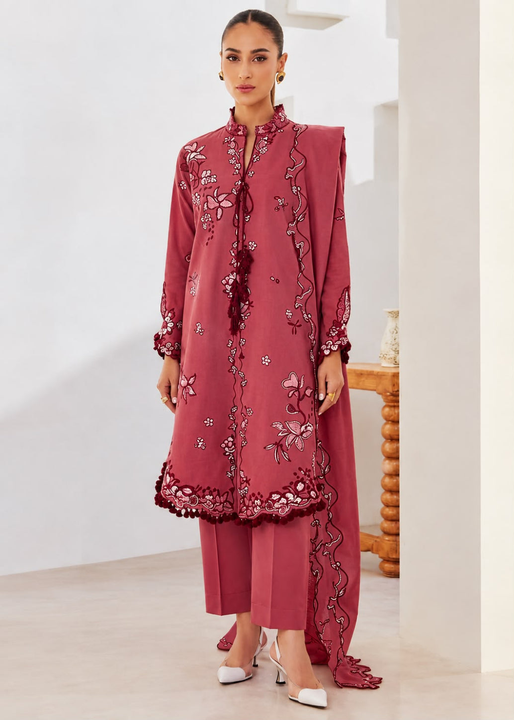 A woman modeling a dusty rose pink ethnic three-piece suit with intricate floral embroidery and border detailing, standing in a bright, modern indoor setting.