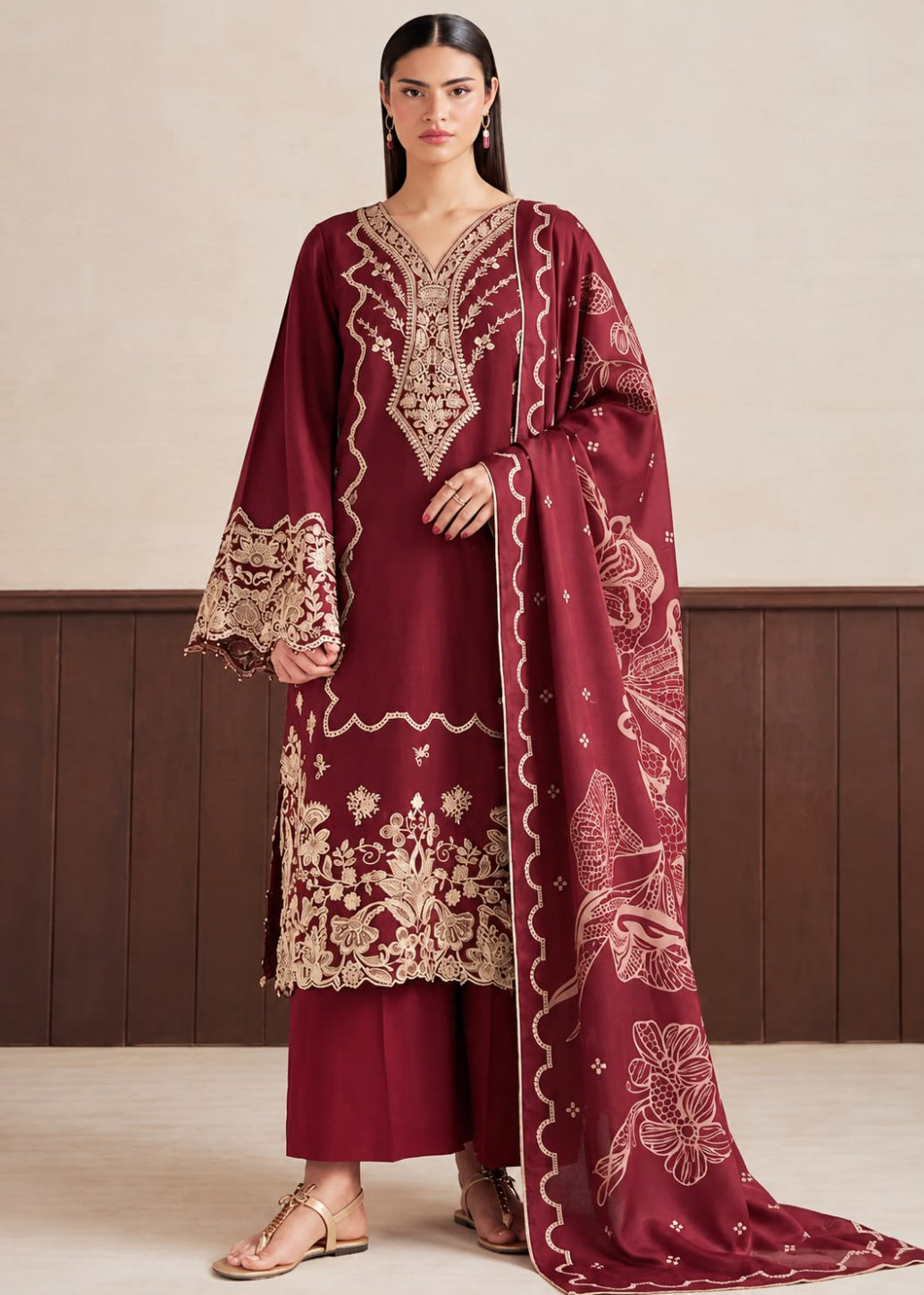 A woman models a traditional Indian salwar kameez suit in deep maroon with intricate beige and gold embroidery. The set includes a long-sleeved tunic with ornate embroidery on the neckline, sleeves, and hem, matching maroon trousers, and a long dupatta featuring floral and bird motifs.