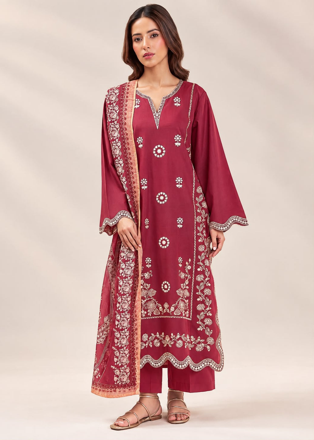 A woman modeling a traditional Indian salwar kameez set in deep maroon, featuring intricate white and gold floral embroidery, a matching dupatta, and matching trousers, set against a neutral beige studio background.