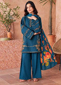 A woman models a teal blue three-piece salwar kameez suit with gold embroidery and a matching floral dupatta in an outdoor patio setting.