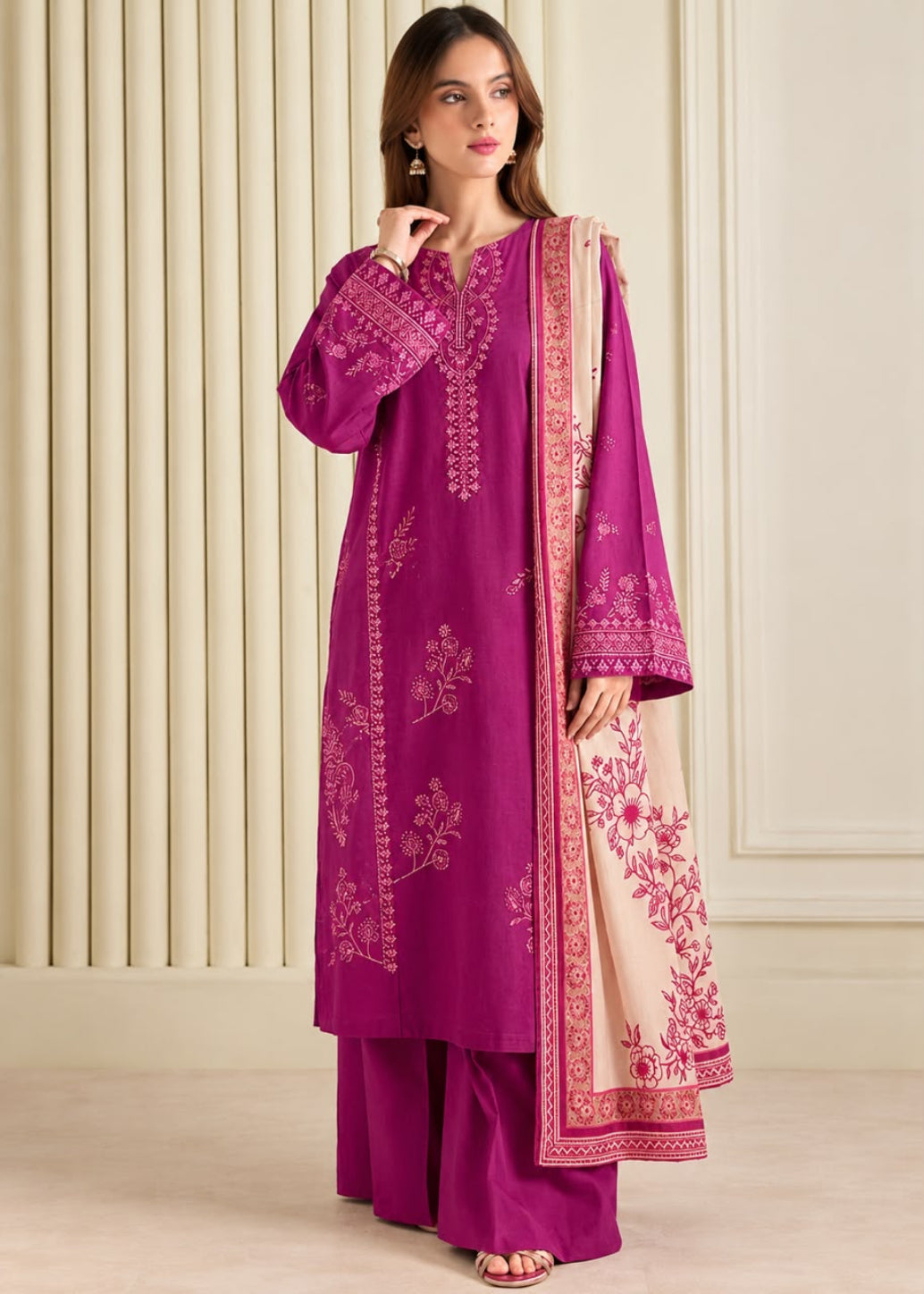 A woman models a traditional Indian salwar kameez suit in magenta with gold embroidery, paired with a beige dupatta featuring pink floral patterns and a gold border, set against a cream paneled background.