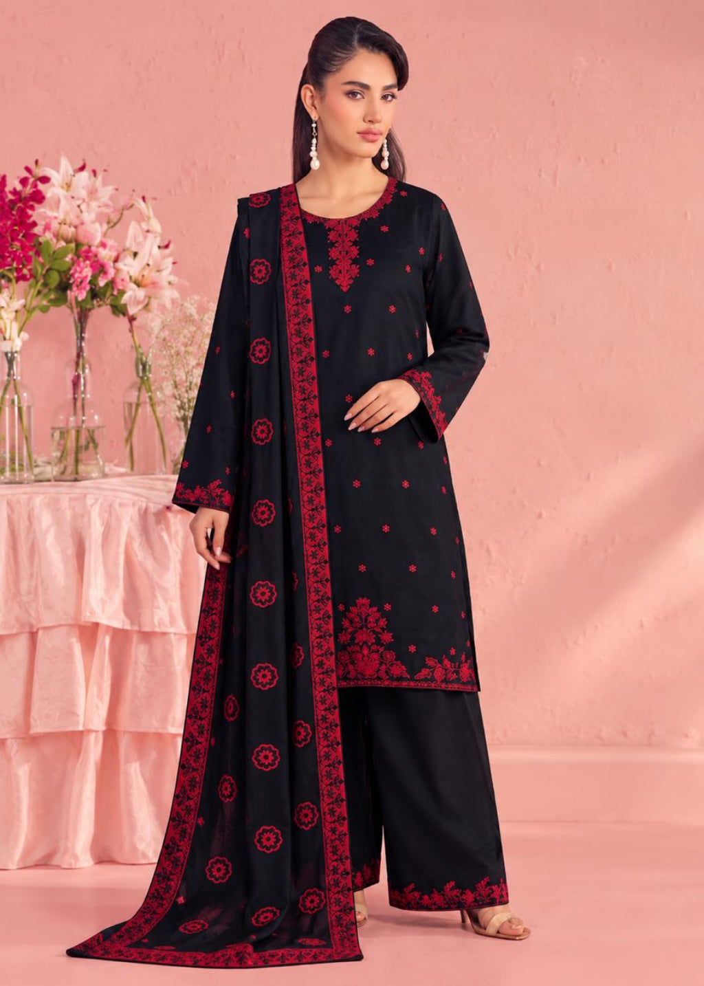 A woman models a black three-piece traditional outfit with red embroidery, featuring a long kurta, wide-leg pants, and a matching dupatta, set against a pink studio backdrop with decorative flowers.