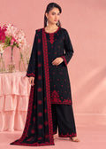 A woman models a black three-piece traditional outfit with red embroidery, featuring a long kurta, wide-leg pants, and a matching dupatta, set against a pink studio backdrop with decorative flowers.