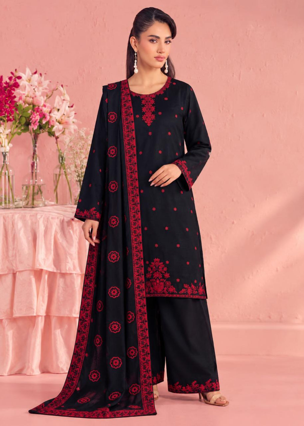 A woman models a black three-piece traditional outfit with red embroidery, featuring a long kurta, wide-leg pants, and a matching dupatta, set against a pink studio backdrop with decorative flowers.