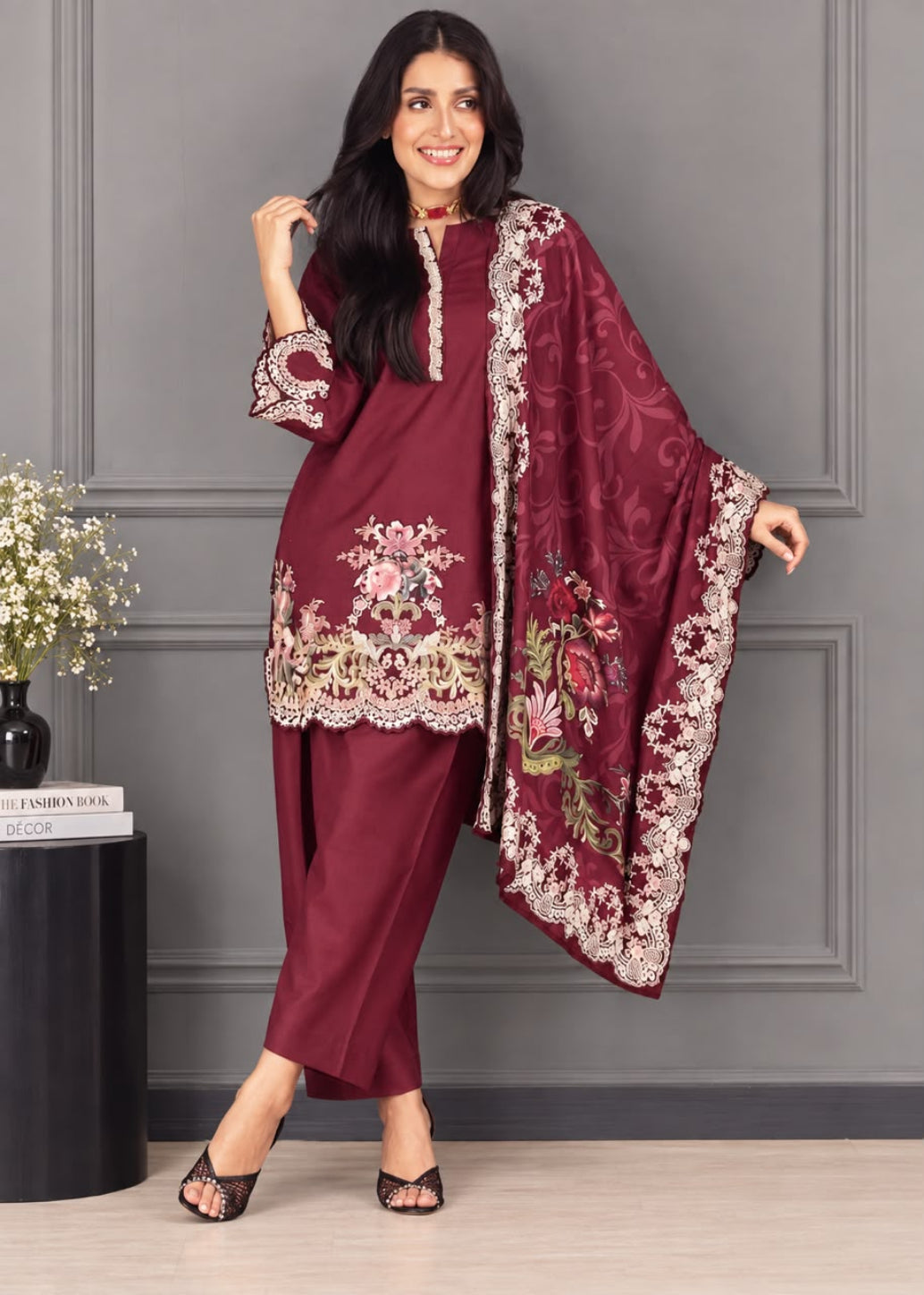 A woman models a three-piece maroon salwar kameez suit with intricate gold and white embroidery, paired with a matching embroidered dupatta, set against a gray paneled wall.