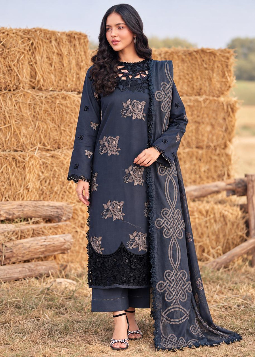 A woman modeling a dark navy blue ethnic suit with beige floral embroidery and a matching dupatta, set against a rustic background of stacked hay bales and a wooden fence.
