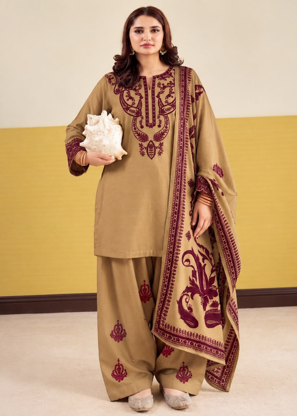 A woman models a traditional three-piece salwar kameez suit in khaki beige with deep maroon embroidery. The set includes a long-sleeve tunic with ornate embroidery on the chest and sleeves, matching wide-leg trousers with small embroidered motifs near the hems, and a coordinating dupatta featuring a large floral print and border design.