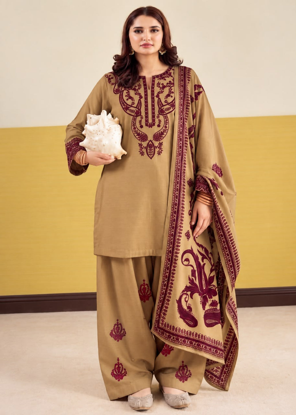 A woman models a traditional three-piece salwar kameez suit in khaki beige with deep maroon embroidery. The set includes a long-sleeve tunic with ornate embroidery on the chest and sleeves, matching wide-leg trousers with small embroidered motifs near the hems, and a coordinating dupatta featuring a large floral print and border design.