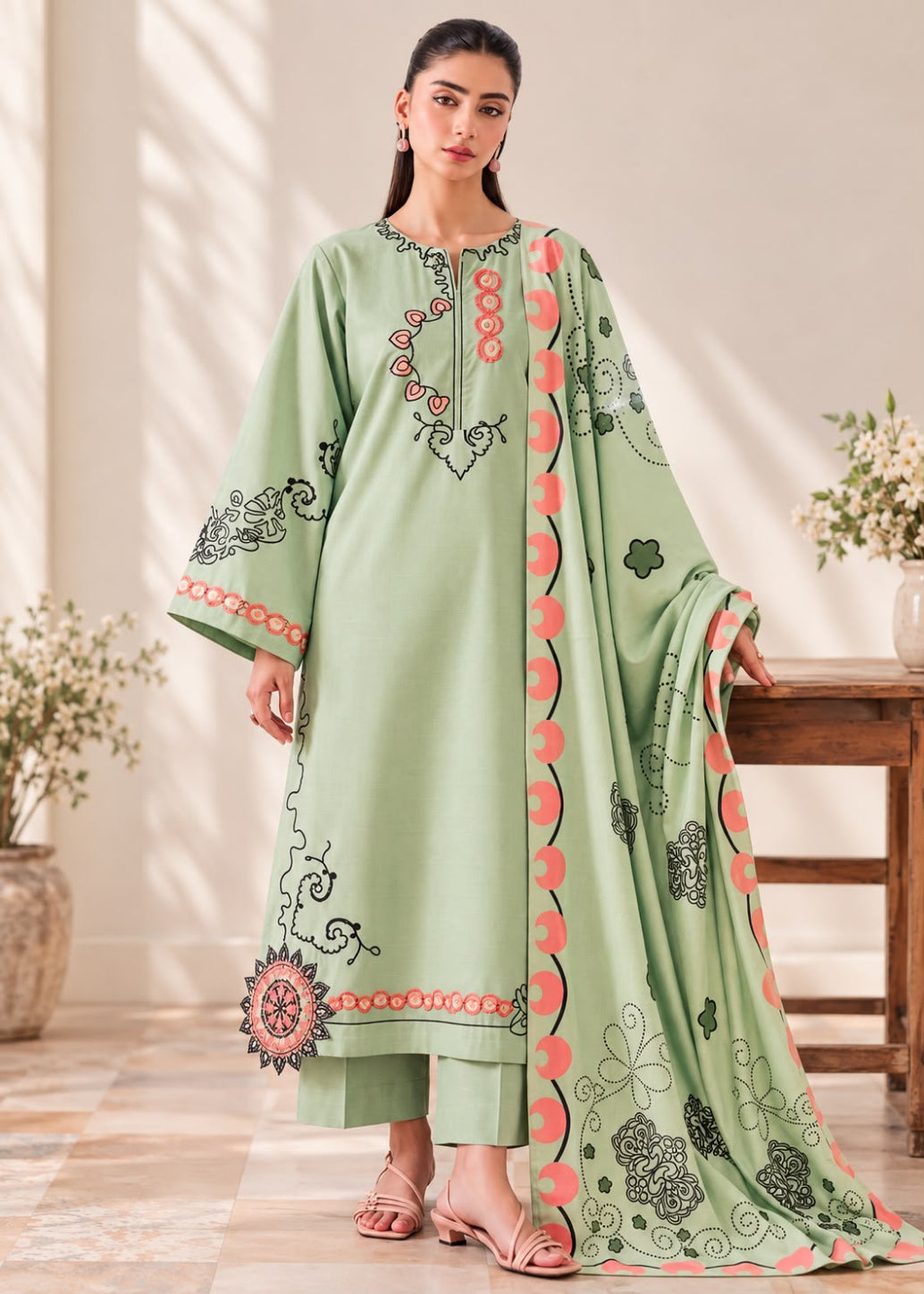 A woman models a light sage green ethnic suit set featuring a long kurta with black and coral embroidery, matching trousers, and a coordinating dupatta with floral patterns, set against a warm-toned indoor background.