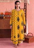 A woman modeling a bright yellow traditional kurta set with black printed motifs, standing in a styled studio setting with a pink textured wall, wooden furniture, and a wicker basket.