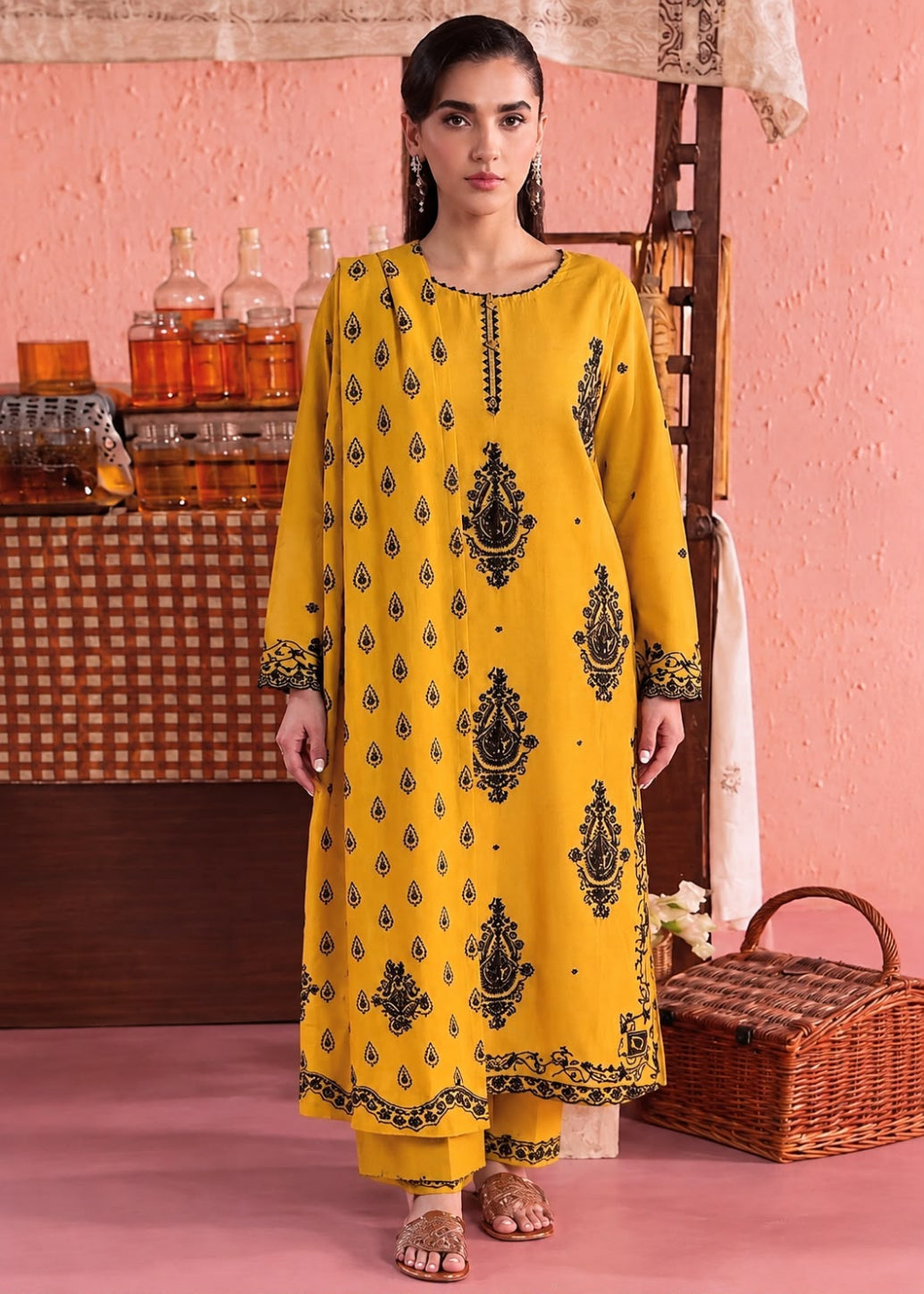 A woman modeling a bright yellow traditional kurta set with black printed motifs, standing in a styled studio setting with a pink textured wall, wooden furniture, and a wicker basket.