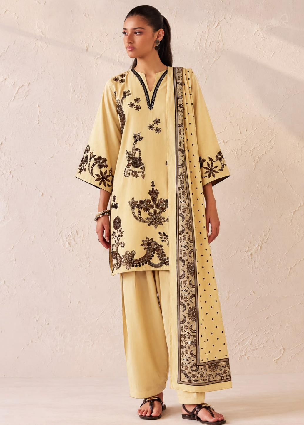 A woman modeling a traditional Indian outfit consisting of a light yellow kurta with black floral embroidery, matching wide-leg trousers, and a coordinating dupatta, set against a textured beige wall.