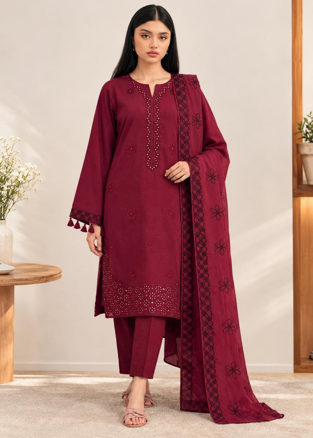 A woman modeling a deep maroon three-piece salwar kameez suit with matching pants and a dupatta, featuring embroidered details and tassel accents, set against a warm beige indoor background.