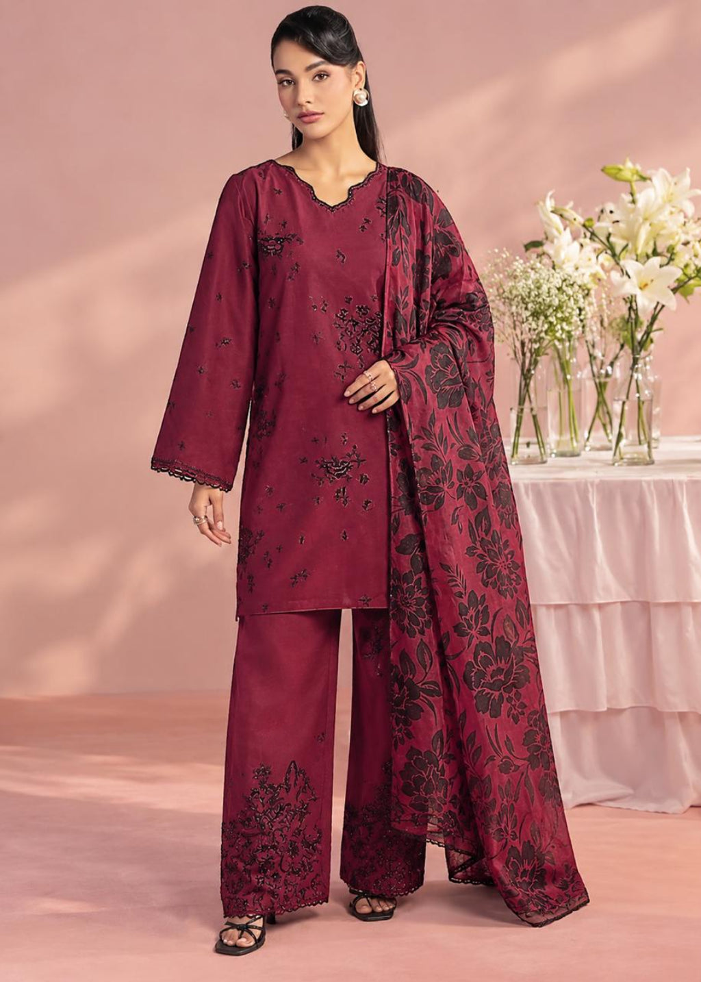 A woman models a three-piece traditional outfit in deep maroon with black floral embroidery, consisting of a long-sleeve kurta, wide-leg trousers, and a matching dupatta, set against a soft pink studio backdrop with a floral table in the background.