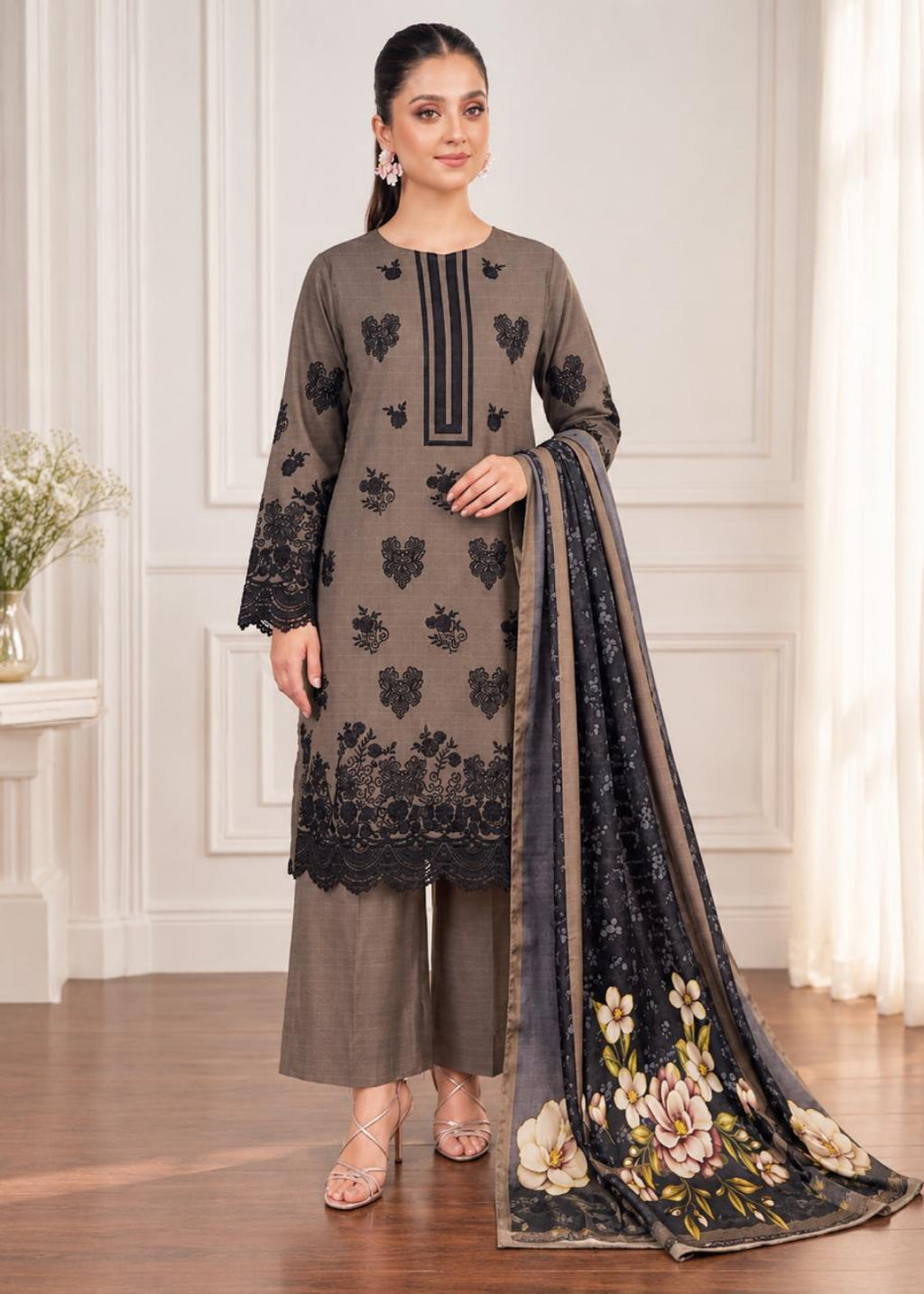 A woman models a three-piece salwar kameez suit in taupe brown with black floral embroidery and a matching printed dupatta, set against an elegant cream-colored interior background.