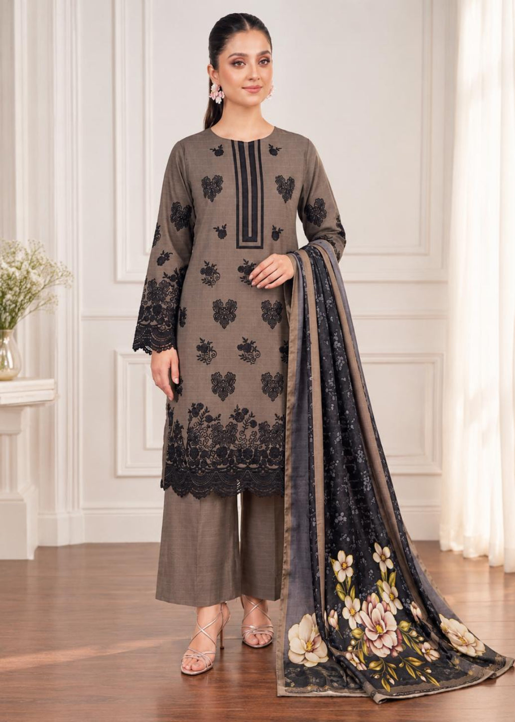 A woman models a three-piece salwar kameez suit in taupe brown with black floral embroidery and a matching printed dupatta, set against an elegant cream-colored interior background.