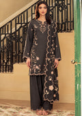 A woman modeling a dark charcoal gray three-piece kurta set with beige floral embroidery, standing in front of a beige wall and black metal staircase railing.